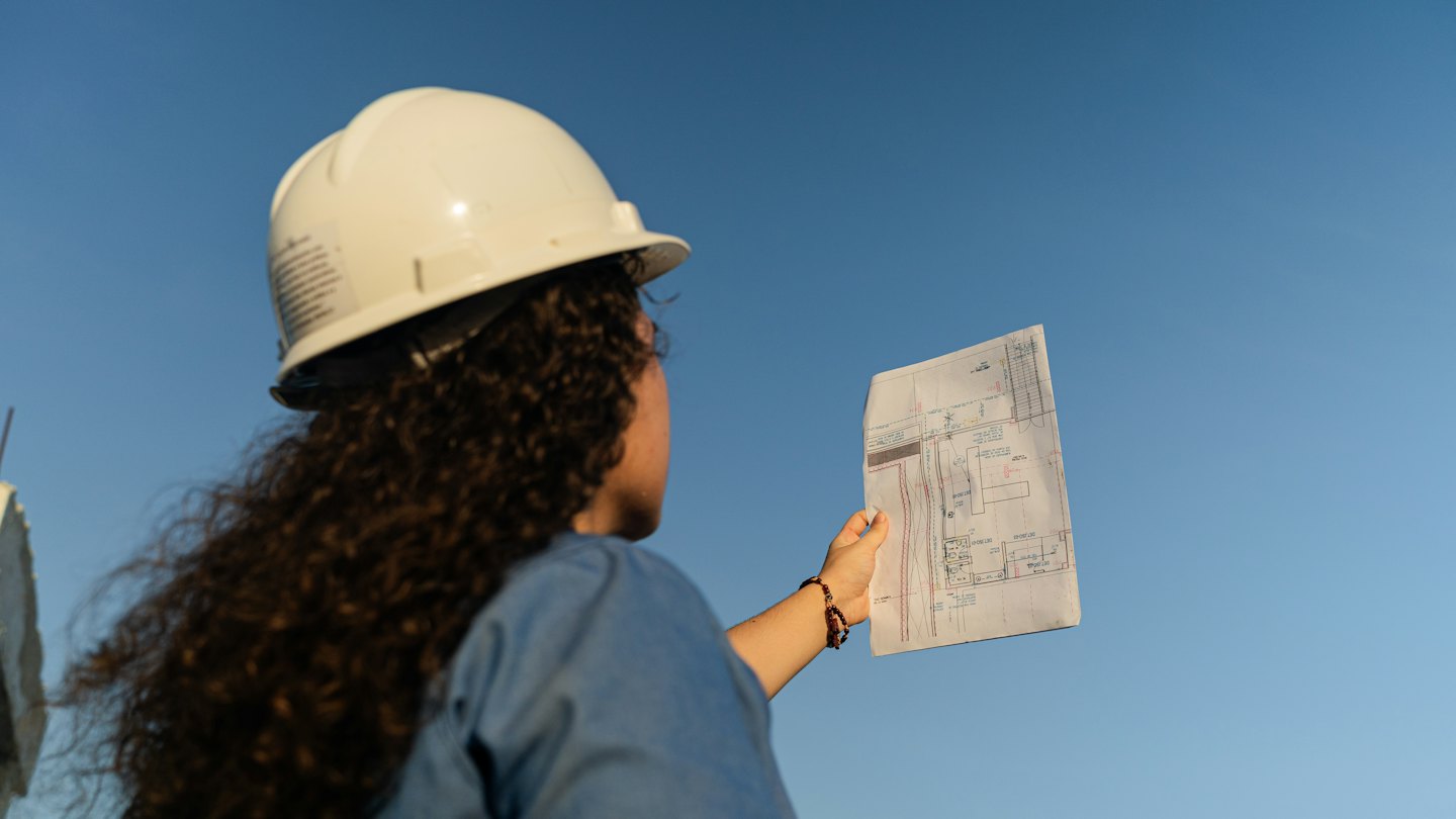 Ingeniero revisa planos arquitectónicos con cielo azul de fondo.